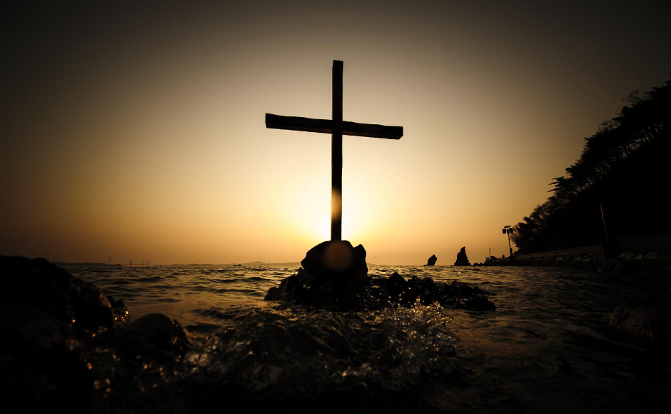 A cross sitting on a rock surrounded by crashing waves at sunset