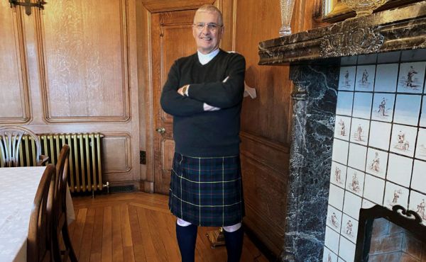 Iain Greenshields wearing a kilt standing beside a fireplace