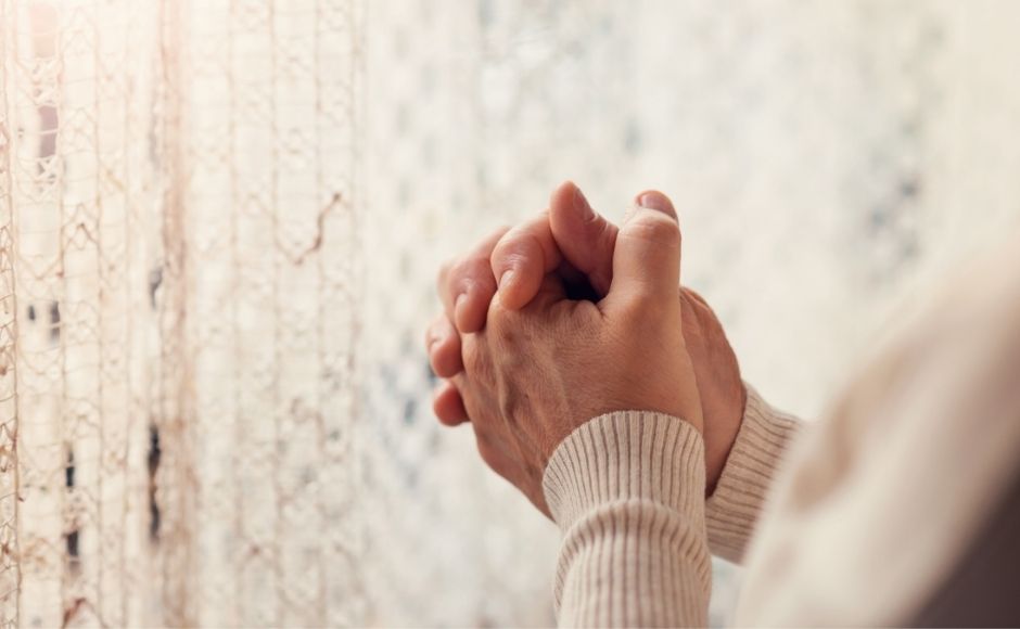 Praying hands standing beside white curtained windows