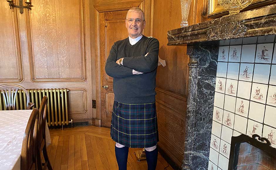 Rt Rev Iain Greenshields wearing a kilt standing beside a fireplace